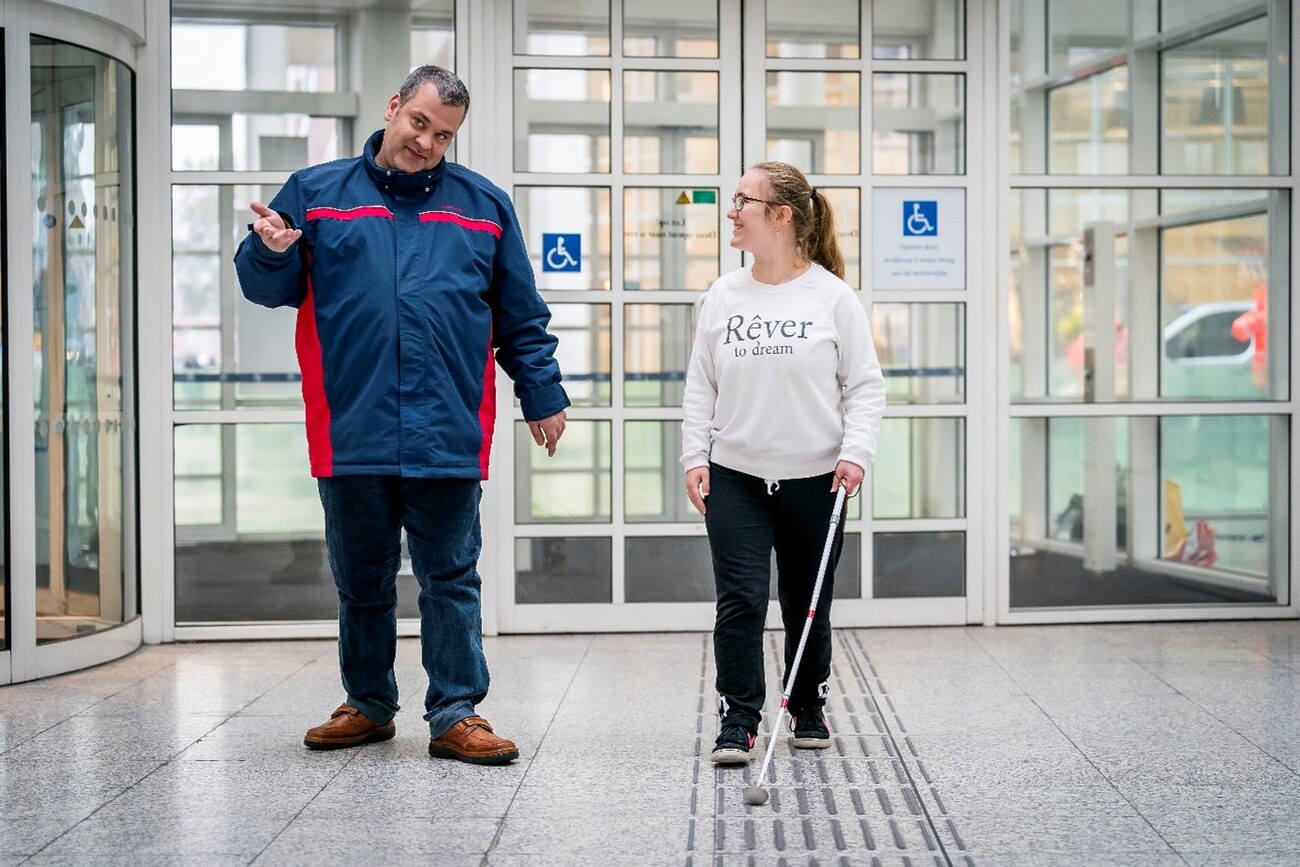 Een man en een vrouw lopen met elkaar door de ingang van het Stadhuis. De man heeft een blauw-rode jas, een spijkerbroek en bruine schoenen aan. De vrouw heeft een witte trui, een zwarte broek en zwarte schoenen aan. Zij zijn met elkaar in gesprek. De vrouw heeft een geleidestok en loopt over een geleidelijn.