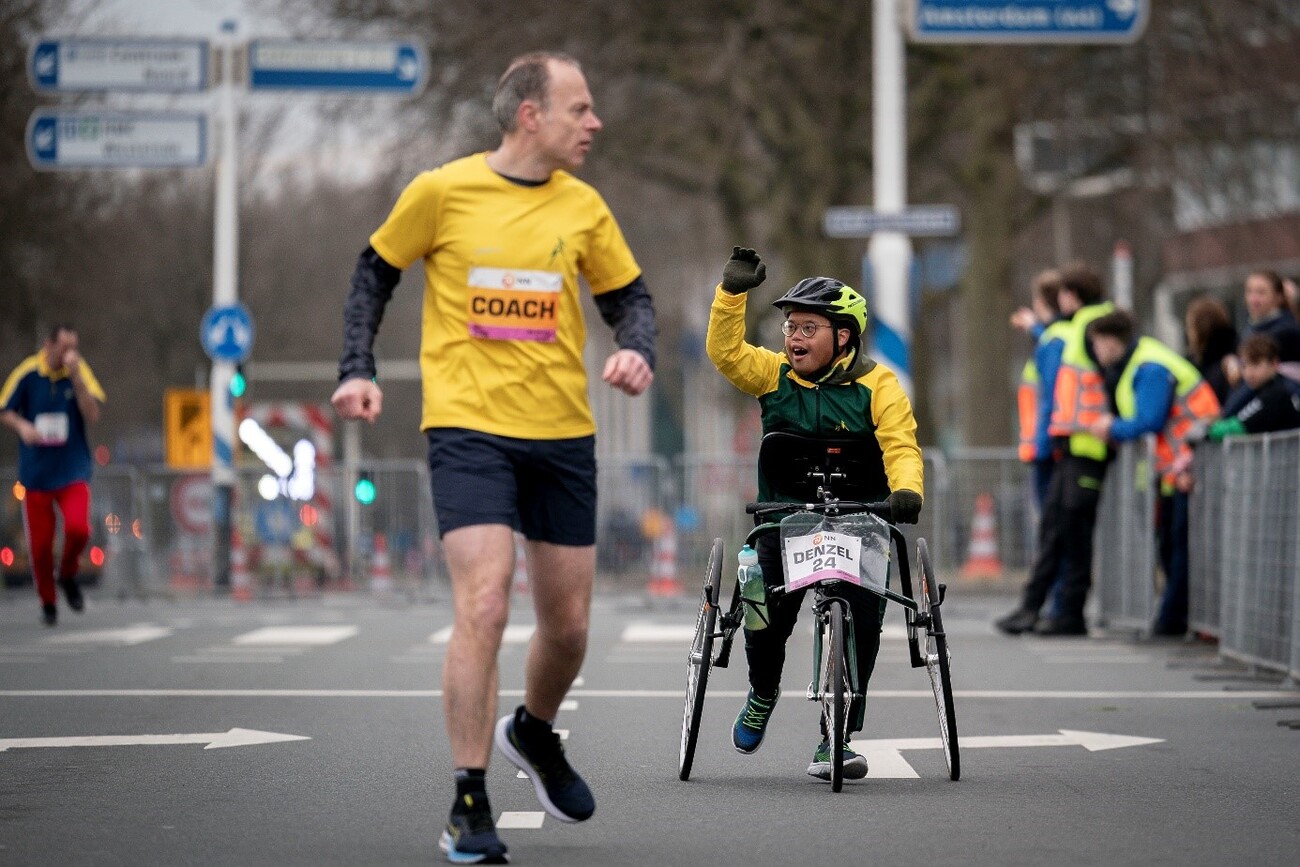 Een man en een jongen doen mee aan hardloopwedstrijd. Zij hebben allebei geel-zwarte sportkleding aan. De jongen heeft een helm op en gebruikt een hulpmiddel tijdens het hardlopen. De jongen zwaait naar het publiek.