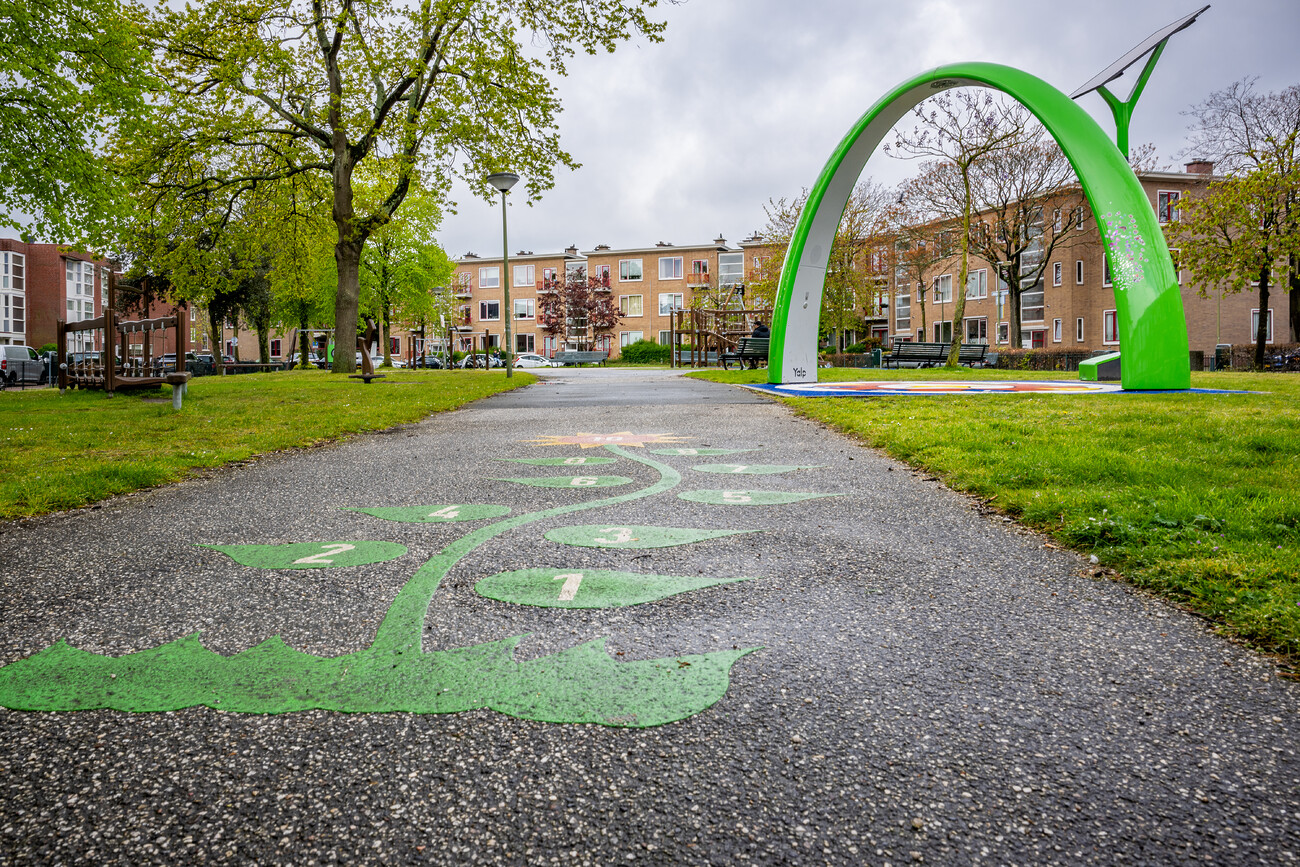 Speeltuin met groen betegeld pad en een groene boog. Omringd door gras en bomen, met flats op de achtergrond.