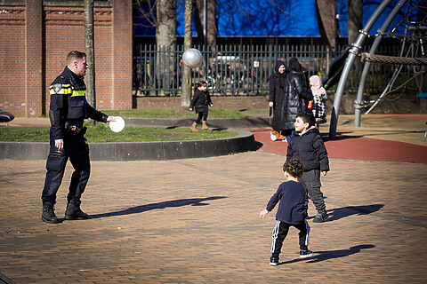 Een politieagent in uniform speelt met 2 jonge kinderen, ongeveer 6 en 3 jaar oud, met een bal.
