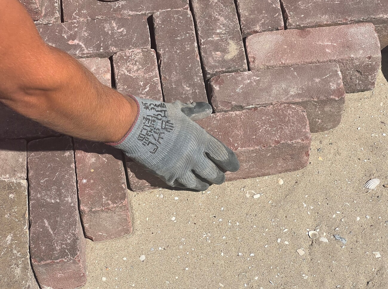 Een close-up foto van een persoon met een grijze handschoen die rechthoekige stoeptegels op zand plaatst.